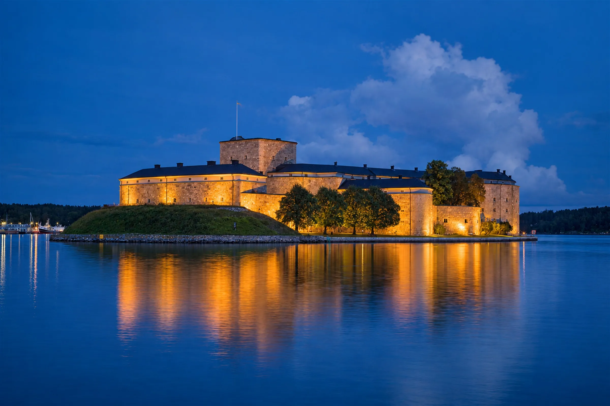 Vaxholms kastell i gult kvällsljus på sin holme, speglat i stilla vatten under blå himmel.