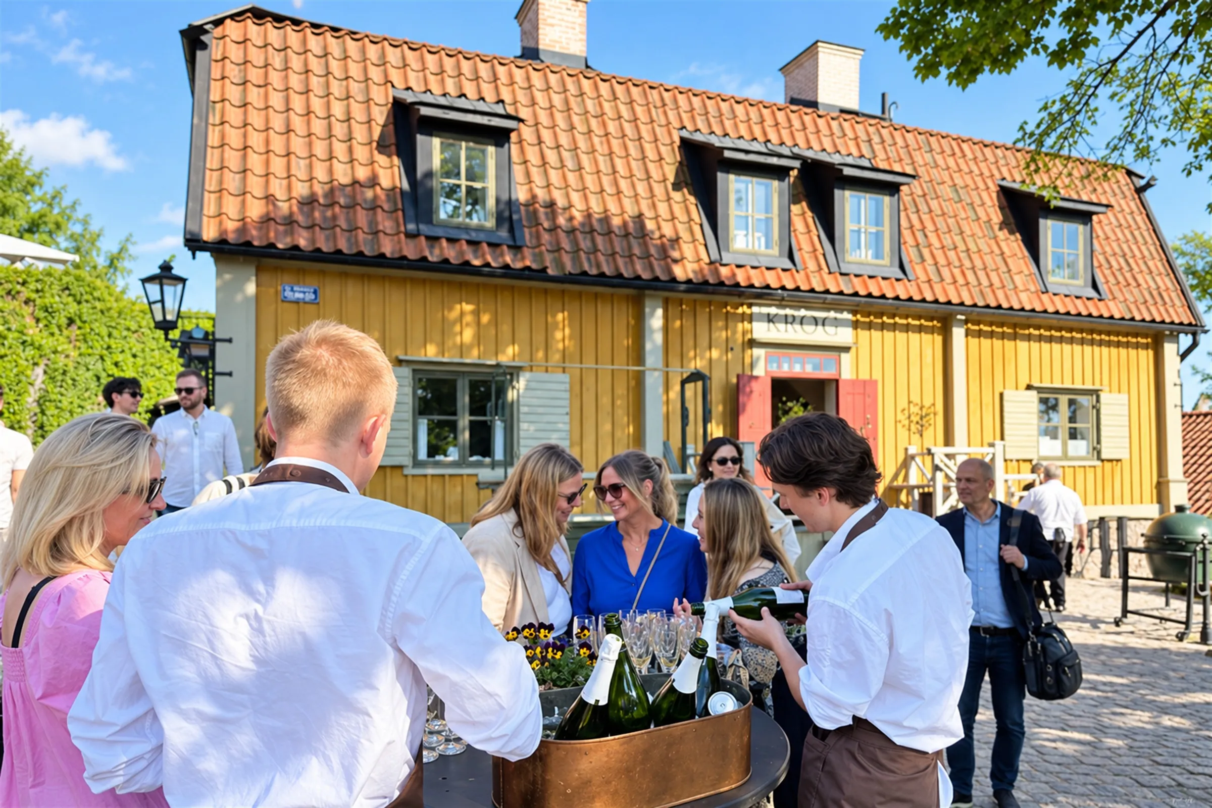 Gäster tar emot ett glas mousserande vin utanför en gul träkrog med tegeltak en solig sommardag.
