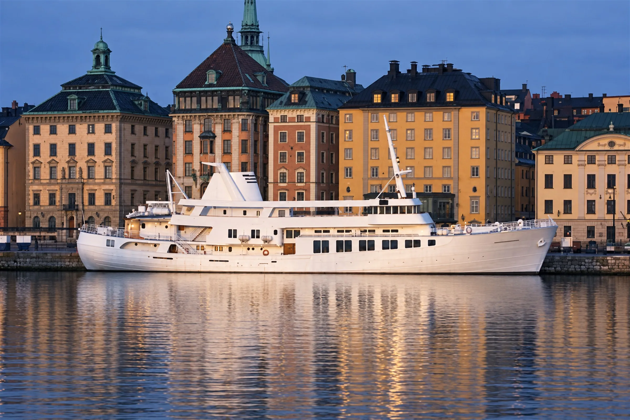 Det vita fartyget Teaterskeppet vid kaj i Stockholm i skymning, med Gamla stans fasader bakom.