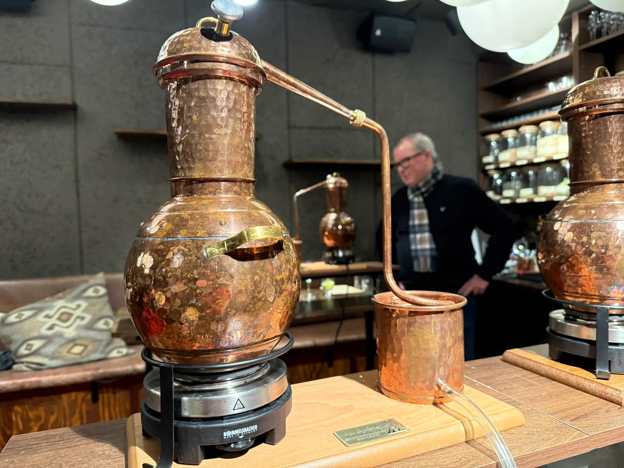 En kopparstill för gindestillering på ett bord i ett rum med hyllor av kryddburkar i bakgrunden.