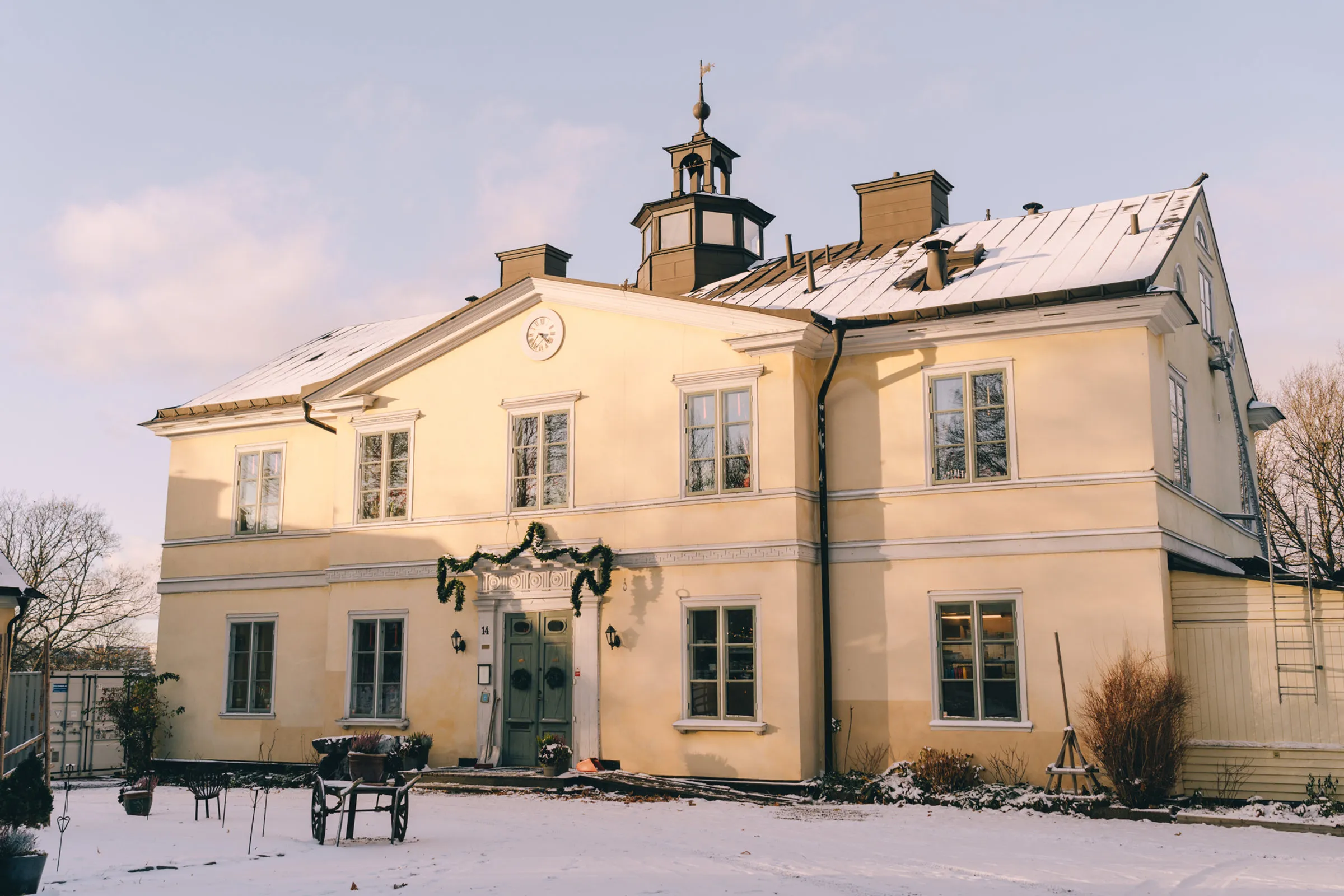 Den gula herrgårdsbyggnaden på Långholmen i snö, med upplysta fönster och en krans på entrén.