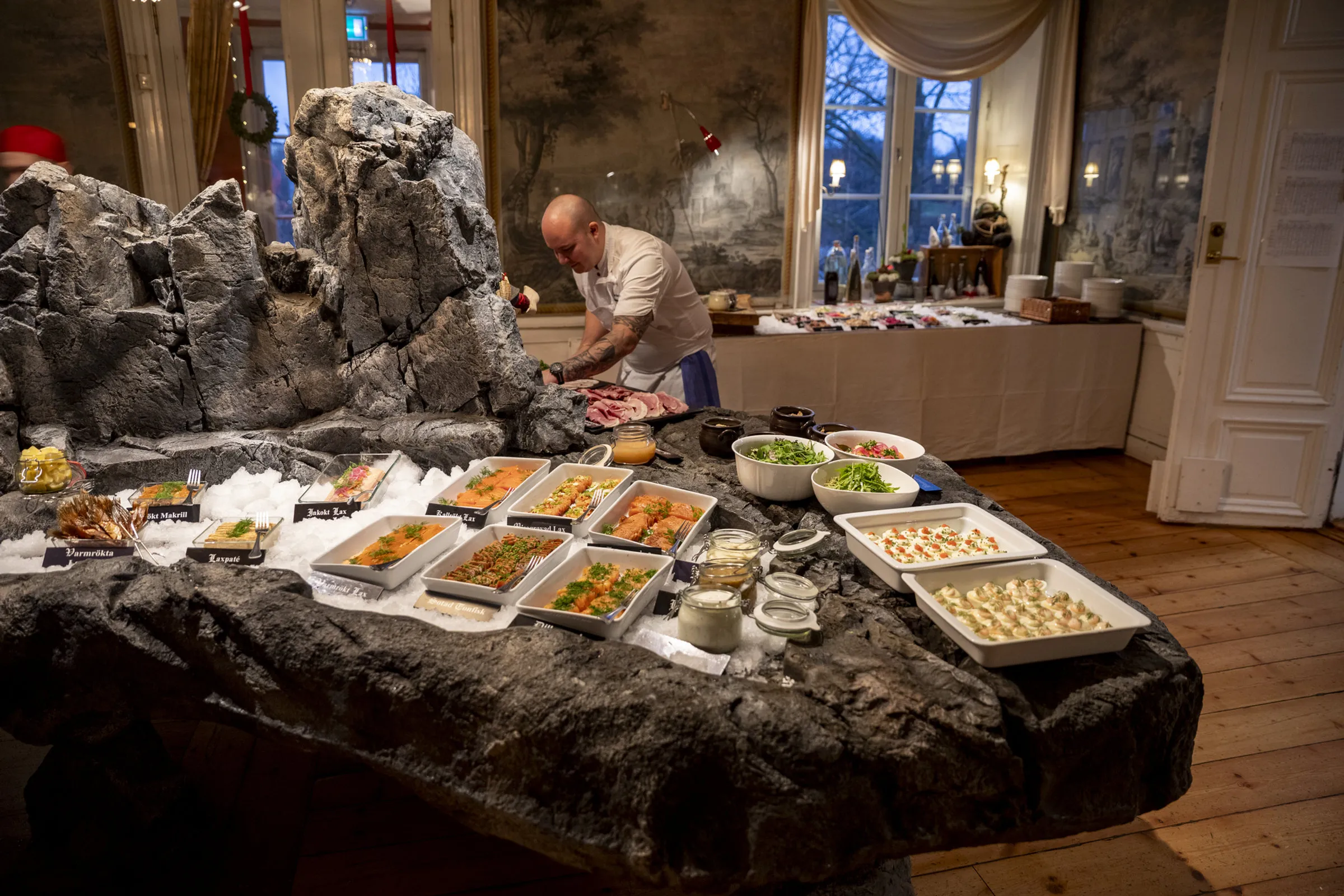 Julbordet uppdukat på ett stenarrangemang med fat av kallskuret, sill och tillbehör, med gäster i bakgrunden.