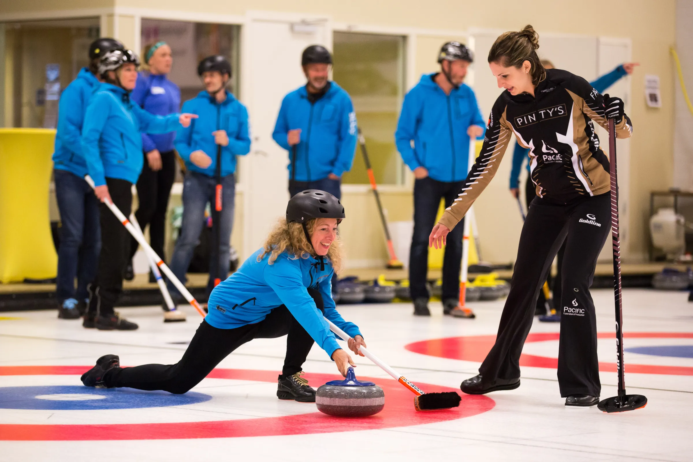 En deltagare i hjälm släpper en curlingsten medan en instruktör visar och kollegor står och tittar på.