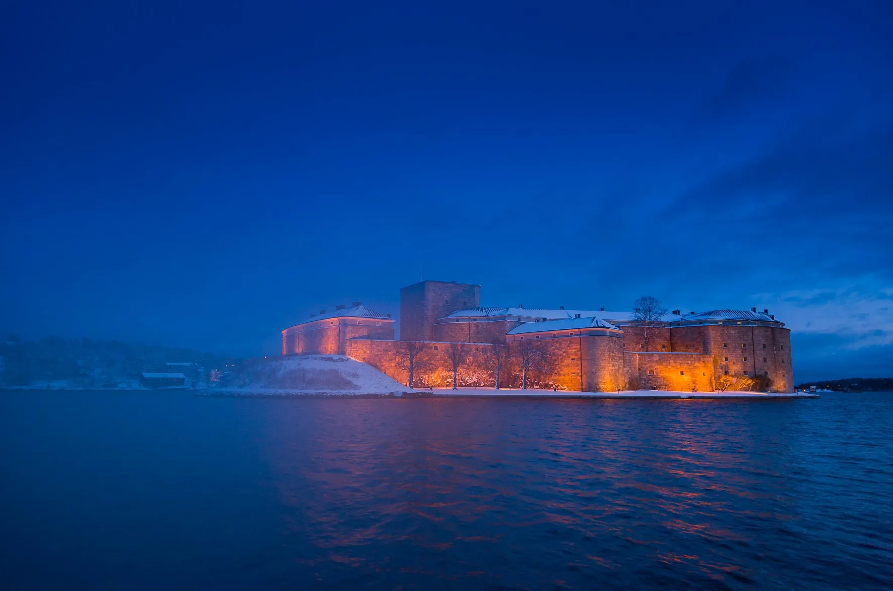 Vaxholms kastell en vinterkväll, den upplysta fästningen i snö med vattnet i förgrunden.