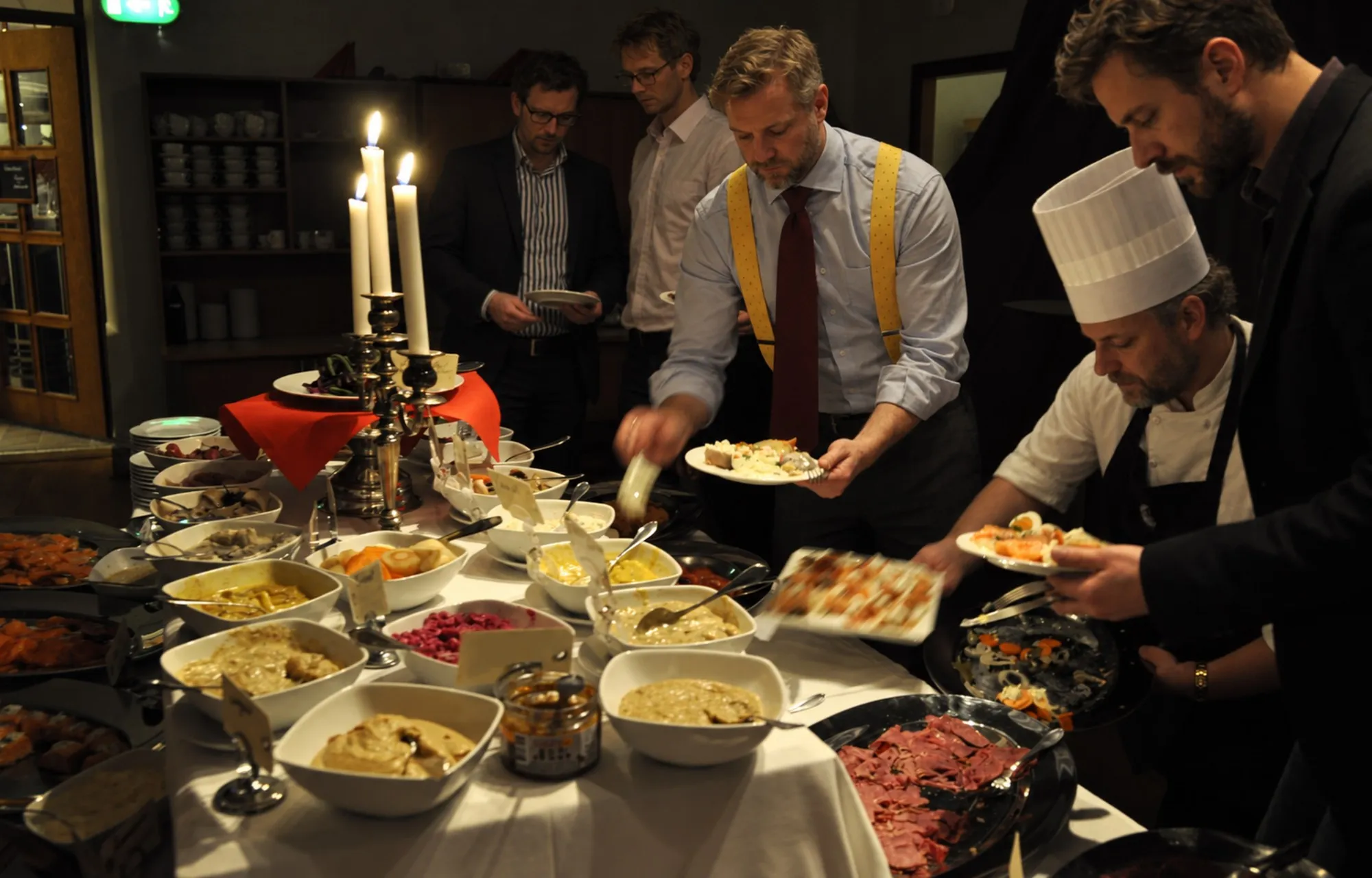 Gäster i kavaj och skjorta tar för sig från ett julbord med sill, lax och skinka i levande ljus, medan en kock i kockmössa fyller på.