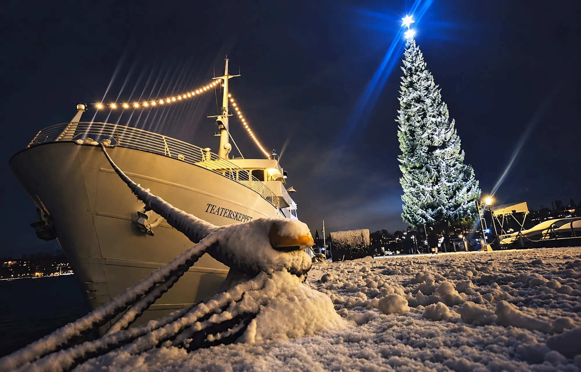 Ett upplyst fartyg förtöjt vid en snötäckt kaj en vinterkväll, med en stor julgran bredvid.