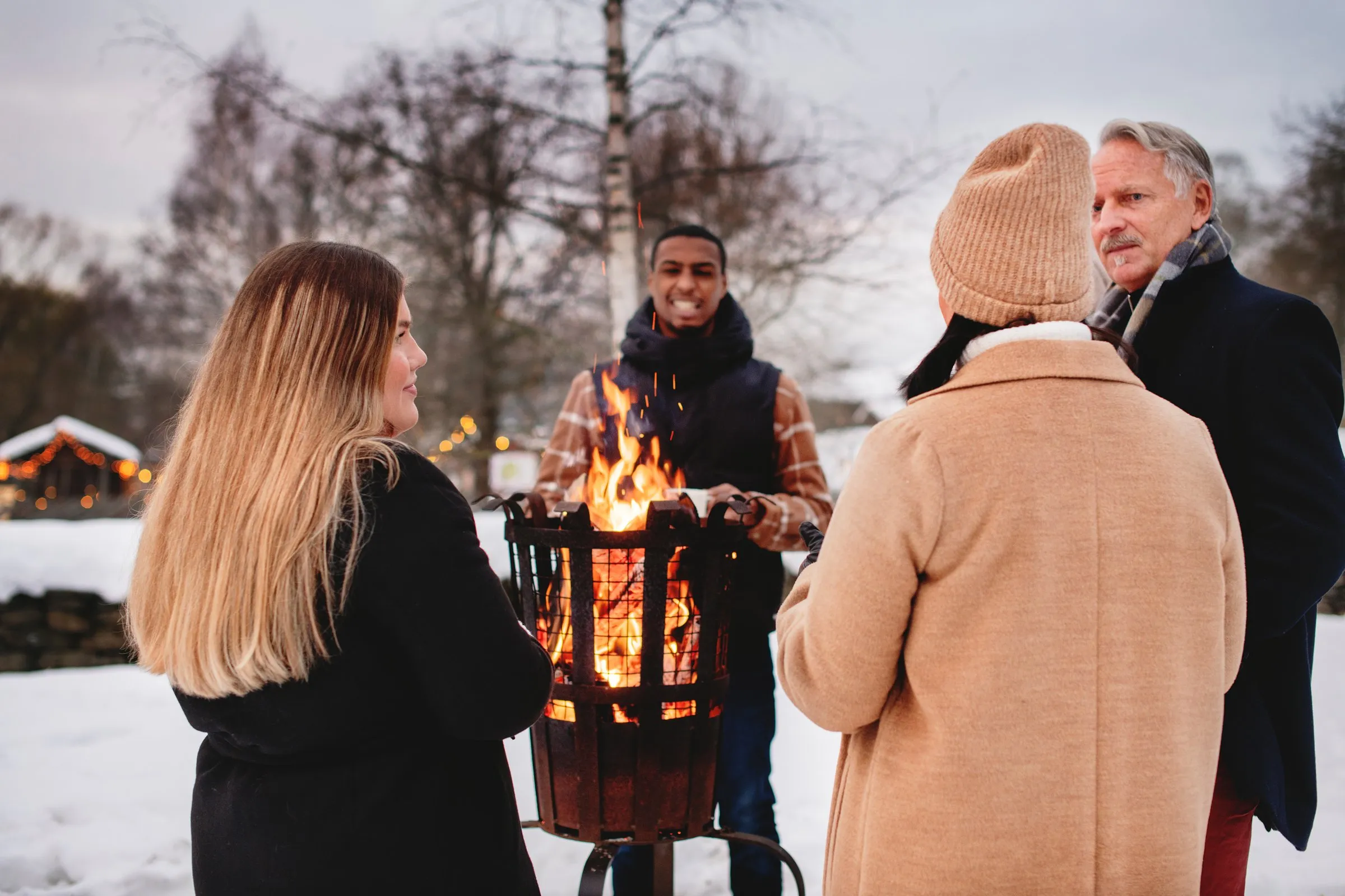 Gäster i vinterkläder samlas kring en vintereld i snön på Skansen, med julbelysta hus i bakgrunden.