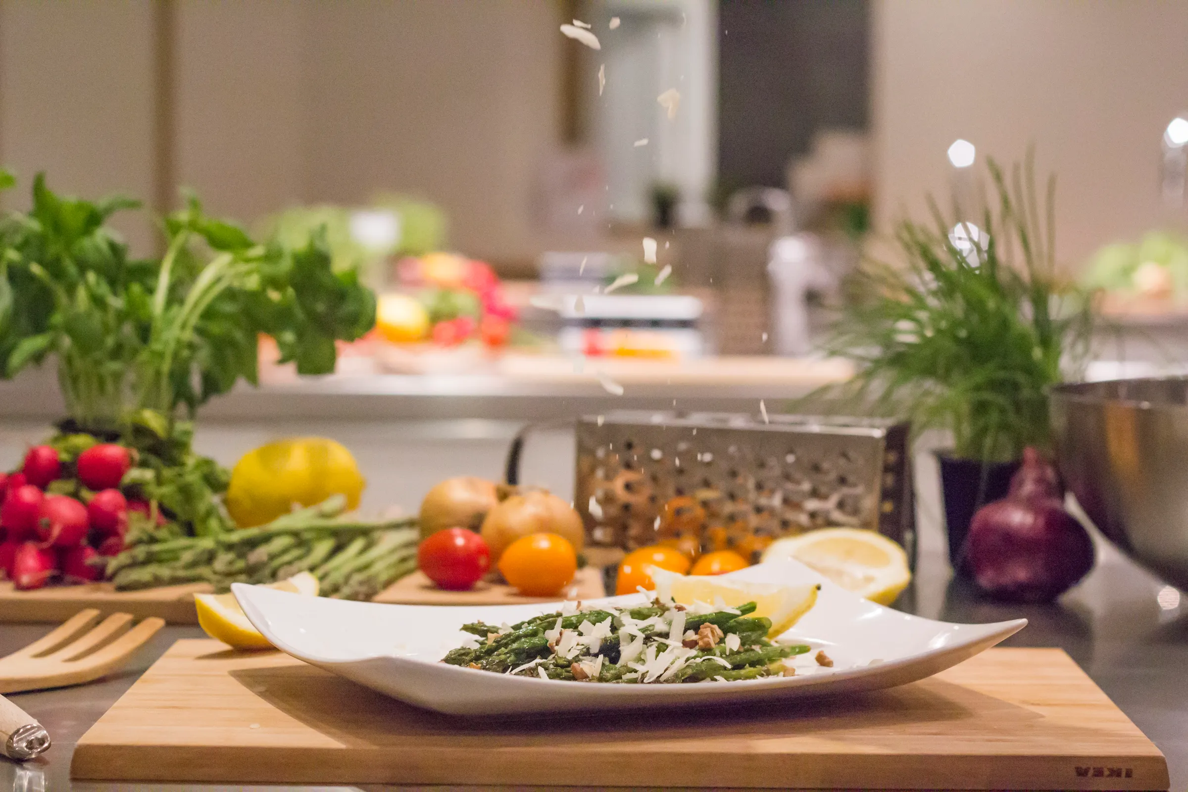 Riven parmesan faller över grön sparris på ett vitt fat omgiven av rädisor, tomater och citron.