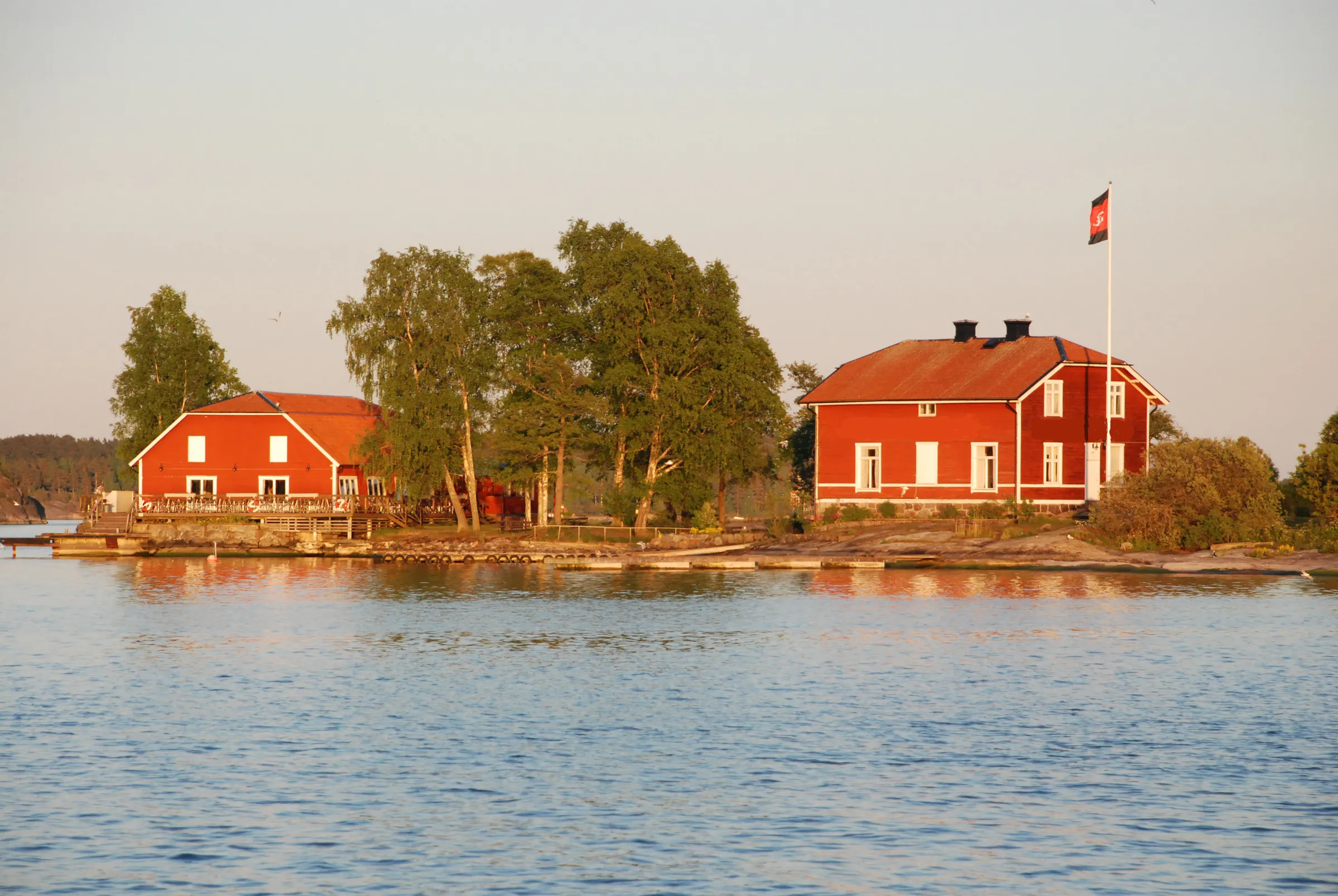 Två röda trähus bland björkar på en klippig holme med flaggstång, sedda från vattnet i kvällssol.