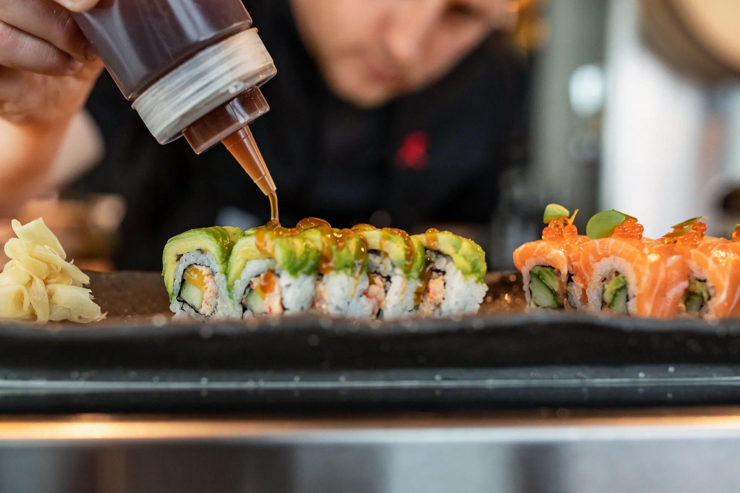 En sushikock ringlar sås över maki med avokado medan inlagd ingefära ligger bredvid på plåten.